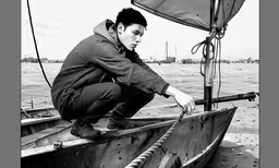 a black and white photo of a man sitting in a boat,a man kneeling on top of a boat in the water