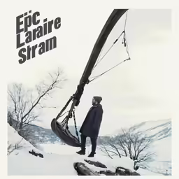 a man in a black coat and jacket stands next to a piece of a boat in the snow