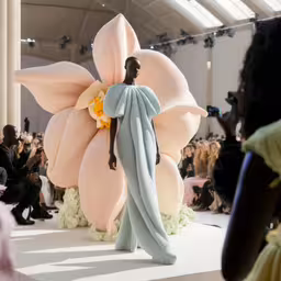 a person in a dress standing on the runway with large flowers,a woman standing on the runway with an enormous flower decoration on display