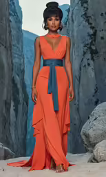 the model in a long, flowing, orange dress stands on a rocky ledge
