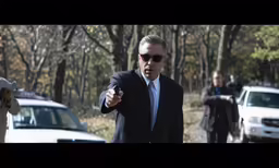 a man in sunglasses and a suit walks behind cars