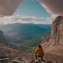 person in yellow jacket and black pants sitting on a rock looking at a mountain range