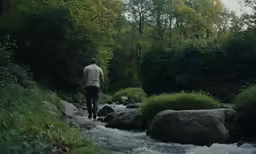 a man walking by a river next to lush green trees