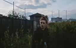 a woman looks up with her hand on her shoulder in front of a fence