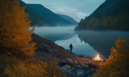 man standing in front of a campfire on the edge of a lake