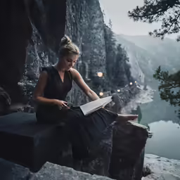 a beautiful young woman reading a book while sitting on a rock by a lake