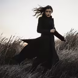 woman in a long black coat standing in tall grasses
