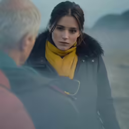 a man with gray hair wearing a jacket and a yellow scarf is looking at a woman on a beach