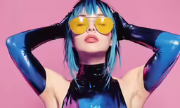 a woman with blue hair, sunglasses and latex outfits poses against a pink background