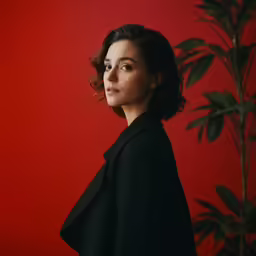 a woman standing against a red wall with leaves and a bush