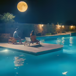two people sitting on wooden chairs next to the side of a swimming pool at night