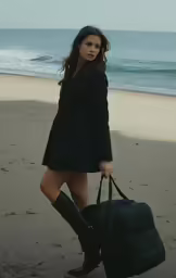 a woman in a coat with a bag and boots on the beach