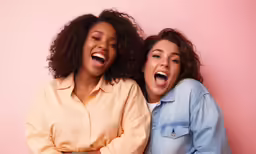 two woman laugh as they stand against a pink background