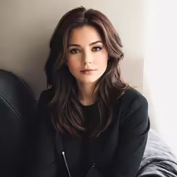 a woman with long hair in a black jacket sits on a couch and leans her head back