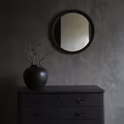 a round mirror above a black dresser with flowers