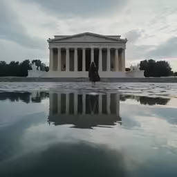 a lone person stands near an ancient building with its reflection in the water