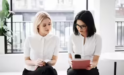 two businesswomen in black pants and white shirt sitting down looking at documents