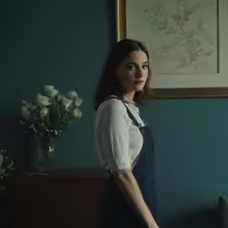 woman in white blouse and blue overalls standing by the windows