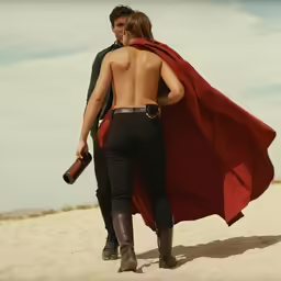 the back of two people with a red cape walking in the sand