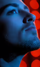 a man with dark eyes and hair looks to the side in front of bright lights