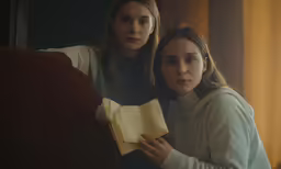 two women stand near each other, one of them holding an open book