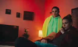 two young women sit in a living room while one of them looks on