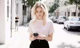 a young woman is standing on a street holding a camera and looking down at her phone