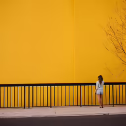 a woman standing on a street corner leaning against a yellow wall