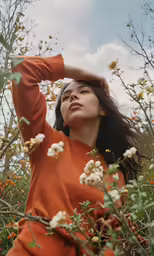 a woman in an orange shirt poses with her arms on her head and the other arm behind her head, surrounded by flowers