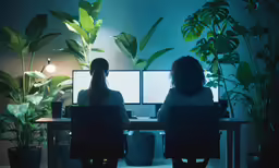 two women are sitting at a desk with two computer monitors