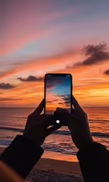 a man holding up an iphone taking a photo on the beach at sunset