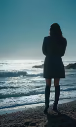 a woman with her back to the camera and head turned to the side stands on the beach with her arms wrapped around herself