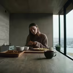 the woman is sitting at the table, she is eating from a bowl