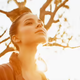 a girl is standing in the sun by a tree