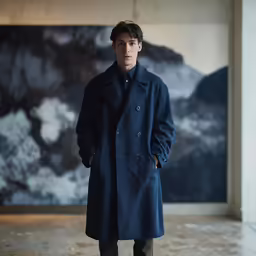 a young man stands wearing a long coat