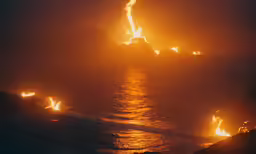 there are people standing on the shoreline looking at a boat with some bright flames