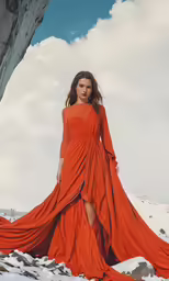 a woman wearing a long red dress poses on a rocky hill