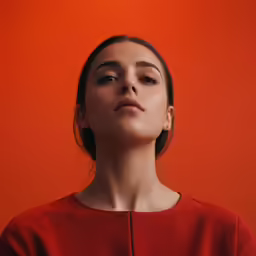 a woman in a red dress stares upward against an orange background