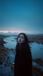 a woman stands on a dirt path by the water in a dark sky