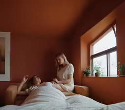 two woman in a bedroom, one sitting on a bed and one standing up