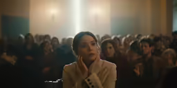 a woman in a white shirt is sitting in a room and looking off into the distance with a large group of people around