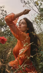 woman with long hair wearing an orange dress sitting on some tall flowers