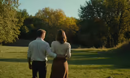 a man and woman walk in the grass with trees in the background
