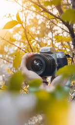 a hand holding up a camera that is in front of some trees
