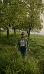 a woman walks through tall grass and trees