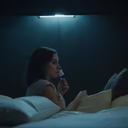 a young girl sits on her bed, with a light shining on her head and looking out a window