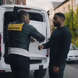two men wearing black are near the back of a van