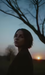 a woman standing next to a tree with the sun behind her