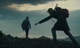 a man holds his backpack with his hands while another man with his back pack walks along the cliff