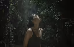 a woman is standing in the middle of the forest, looking up into a sky with light